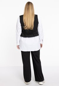 Schwarze Nadelstreifen-Weste über einem weißen Hemd mit Knöpfen, kombiniert mit schwarzen Nadelstreifen-Hosen und weißen Sneakers, von hinten gesehen.