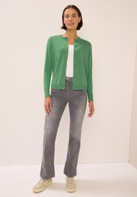Zelený z button-up kardigan přes bílý top, spárovaný s lehkými šedými zvonovými džínami a béžovými teniskami. Hladká textura, jednoduchý design.