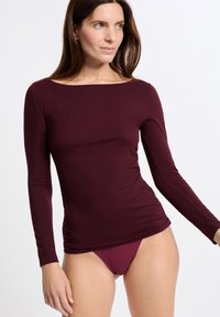 Donna che indossa un top a maniche lunghe attillato color bordeaux e biancheria intima abbinata, in piedi contro uno sfondo semplice e chiaro, con lo sguardo rivolto di lato.