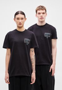 Czarne bawełniane koszulki z logo na przodzie, okrągłym dekoltem, krótkimi rękawami i luźnym krojem. Noszone przez dwie osoby, z których jedna ma tatuaże.