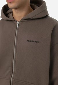 Sudadera con cremallera de color marrón, hecha de una tela suave. Cuenta con capucha, bolsillos laterales y un pequeño logo negro en el pecho. La cremallera es metálica.