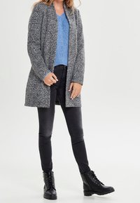 Kvinna klädd i en grå texturerad kappa, blå stickad tröja, svarta skinny jeans och svarta snörkängor, som står mot en enkel bakgrund.