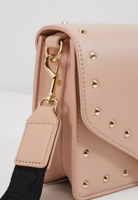 Blush pink leather handbag with gold rivets, featuring a detachable black strap and a lobster clasp. Smooth texture and structured design.