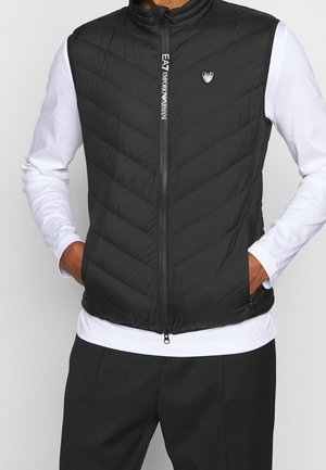 Chaleco negro acolchado sin mangas con cremallera y logo, llevado sobre una camisa blanca de manga larga y pantalones negros, con las manos en los bolsillos del chaleco.