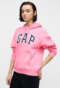 Νεαρός με κοντά σκούρα μαλλιά, που φορεί ένα φωτεινό ροζ φούτερ με κουκούλα και μια μπροστινή τσέπη, με τα γράμματα "GAP", στέκεται με το ένα χέρι πίσω από την πλάτη.