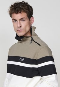 Štýlová mikina s vysokým golierom, khaki vrchom a horizontálnymi pruhmi v čiernej a bielej farbe. Obsahuje zips a vyšívané logo.