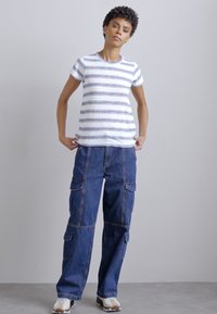 Tricou alb cu mânecă scurtă și dungi albastre și violet, purtat cu pantaloni cargo din denim bleu închis, largi, care au multiple buzunare.