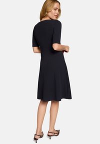 Femme en robe noire mi-longue avec manches au coude, debout et regardant par-dessus son épaule, portant des escarpins slingback marron.