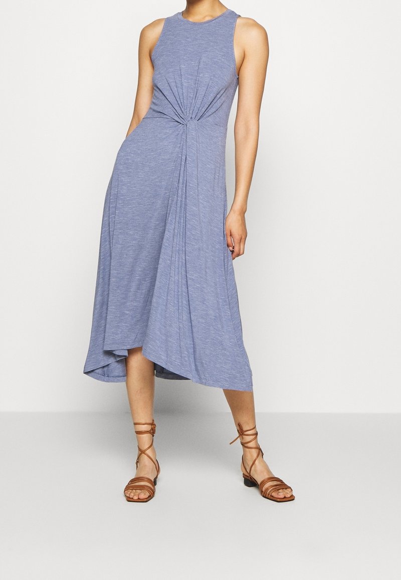 Femme portant une robe midi sans manches bleu clair avec un détail noué froncé à la taille et des sandales plates marron à lacets.