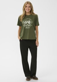 T-shirt en coton vert olive avec un imprimé graphique blanc représentant des ailes et du texte, associé à un pantalon noir ajusté et des chaussures à lacets marron.