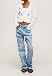 Biały krótki kardigan z wzorem diamentowym i czerwonym kwiatowym haftem, zestawiony z jasnoniebieskimi dżinsami z wysokim stanem i czarnymi sandałami.