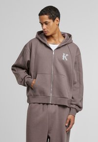 Felpa con cappuccio e zip di colore taupe, realizzata in tessuto morbido. Presenta tasche a marsupio e un logo sottile sul petto. Vestibilità ampia e rilassata.
