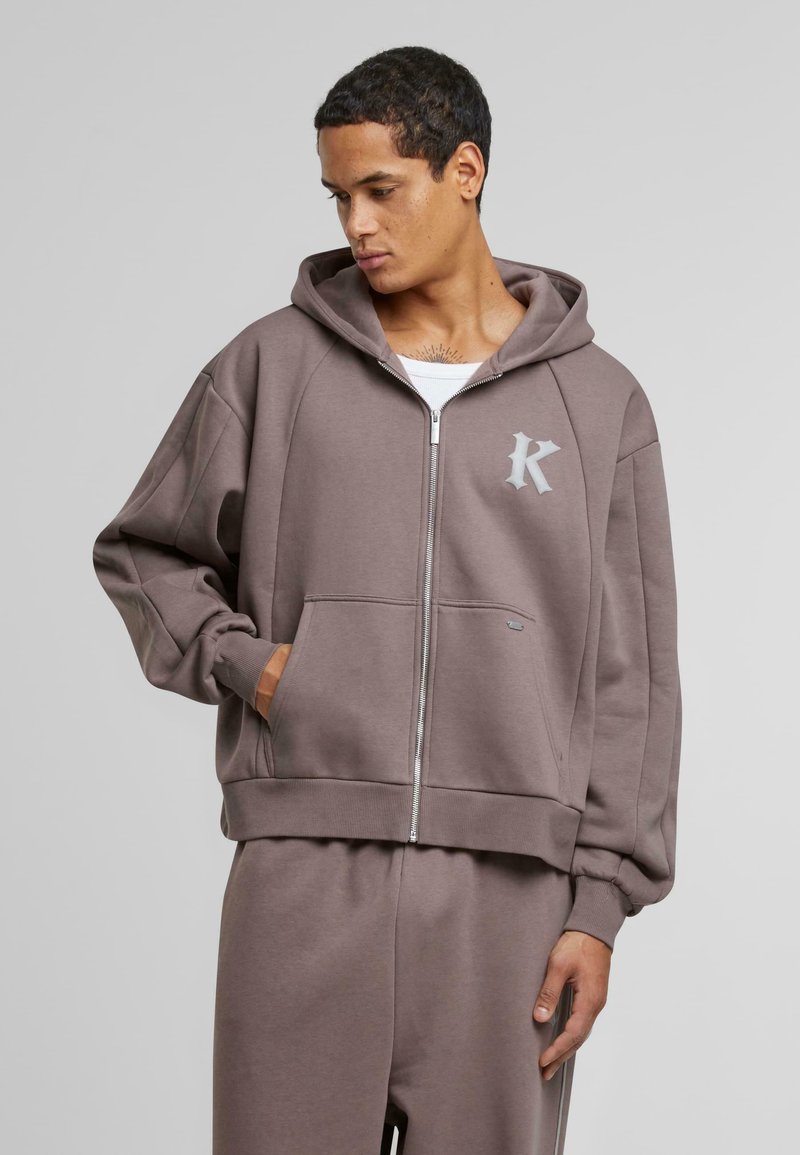 Felpa con cappuccio e zip di colore taupe, realizzata in tessuto morbido. Presenta tasche a marsupio e un logo sottile sul petto. Vestibilità ampia e rilassata.