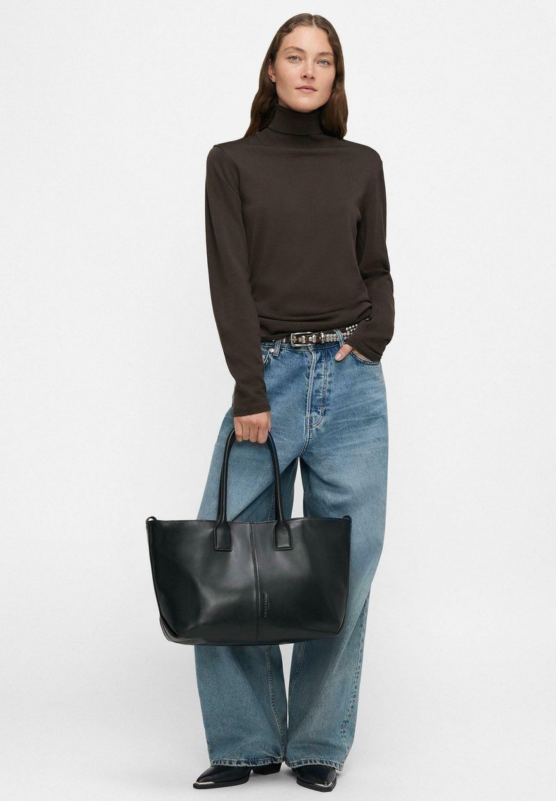 Sac fourre-tout en cuir noir avec deux poignées plates, de grande taille et texture lisse. Porté avec un col roulé marron et un jean bleu clair à coupe large.