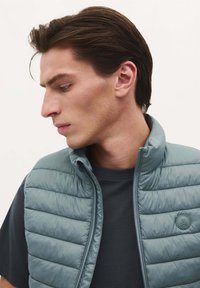 Lichtblauwe puffer vest met een gestikt ontwerp, staande kraag, ritssluiting aan de voorkant en een logo op de patch. Gedragen over een marineblauw T-shirt, profielweergave van dichtbij.
