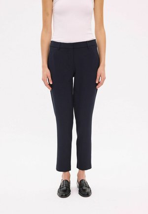 Žena nosí tmavomodré slim fit krátke nohavice, biely tielkový top a čierne kožené mokasíny, stojí na jednoduchom pozadí.