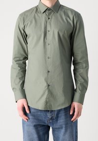 Langsleeve olijfgroene shirt met een knoopsluiting aan de voorkant, gestructureerde kraag en subtiele glans, gecombineerd met blauwe jeans.