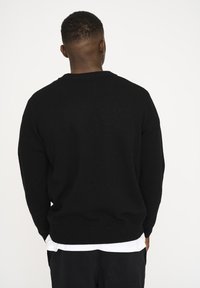 Czarny sweter dzianinowy z okrągłym dekoltem i ściągaczami. Nałożony na biały shirt, o luźnym kroju i teksturowanej tkaninie.