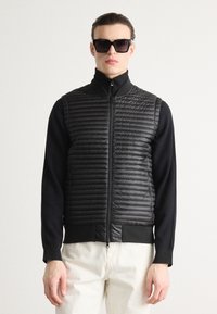 Μαύρο επενδυμένο γιλέκο με φερμουάρ, οριζόντιες ραφές και ψηλό γιακά. Συνδυάστε το με ένα εφαρμοστό μαύρο μακρυμάνικο μπλούζα και λευκά παντελόνια.