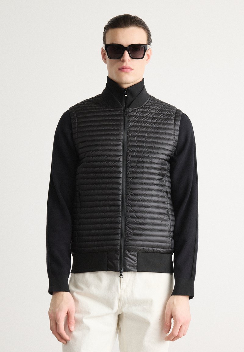 Μαύρο επενδυμένο γιλέκο με φερμουάρ, οριζόντιες ραφές και ψηλό γιακά. Συνδυάστε το με ένα εφαρμοστό μαύρο μακρυμάνικο μπλούζα και λευκά παντελόνια.