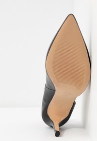 Chaussure à talon haut en cuir noir à bout pointu vue de la semelle avec une semelle beige et un petit talon, positionnée sur un fond blanc.