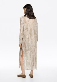Vestido largo de un tono beige claro con un patrón texturizado, mangas largas, aberturas laterales y un dobladillo asimétrico. Lleva unos zapatos planos marrones.