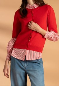 Czerwony dzianinowy kardigan z krótkimi rękawami, ozdobiony złotymi guzikami, nałożony na biało-czerwony w paski T-shirt. Luźne niebieskie jeansy.