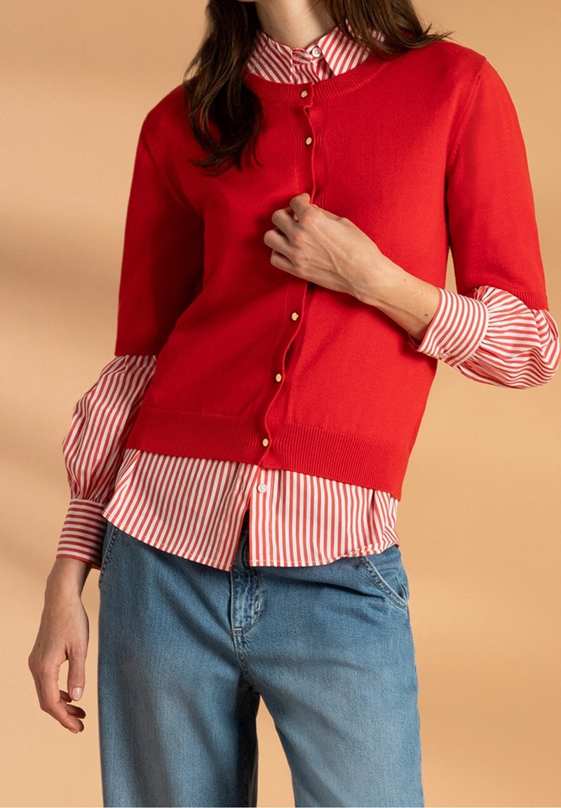Czerwony dzianinowy kardigan z krótkimi rękawami, ozdobiony złotymi guzikami, nałożony na biało-czerwony w paski T-shirt. Luźne niebieskie jeansy.