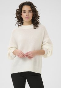 Pulover alb din tricot cu guler înalt, mâneci largi și o textură moale, asortat cu pantaloni negri ajustați.