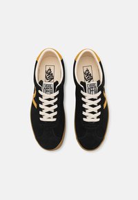 Zapatillas de ante negro con cordones crema, franja lateral amarilla y suela de goma. Presentan un diseño clásico de corte bajo y la marca Vans en el interior.
