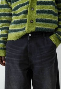 Zielona, w paski, puszysta kamizelka z okrągłym dekoltem i ciemnymi guzikami, w połączeniu z ciemnoniebieskimi spodniami dżinsowymi z przednią kieszenią i srebrnym guzikiem.