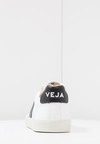 White sneaker with black heel tab, featuring "VEJA" logo. Smooth leather material with a classic low-top design and rubber sole.