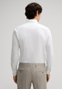 Camicia bianca a maniche lunghe con polsini a bottone, abbinata a pantaloni eleganti beige chiaro con tasche posteriori e dettagli di cucitura discreti.