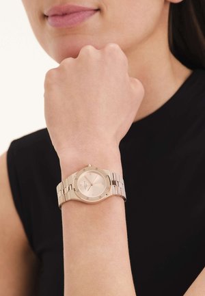 Muñeca con un reloj de metal dorado que marca las 10:11, usado por una persona con una camiseta negra sin mangas y una leve sonrisa.