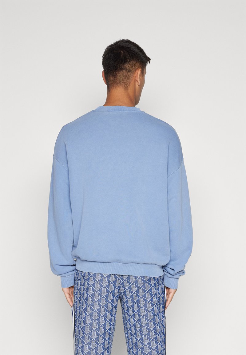 Hombre de espaldas vestido con un suéter grande de color azul claro y pantalones azules con un diseño geométrico, todo sobre un fondo blanco liso.