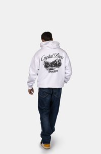 Felpa bianca con cappuccio, caratterizzata da una grande stampa nera di montagne e testo sul retro, abbinata a jeans di denim blu e sneakers.