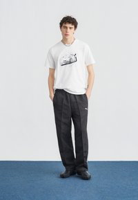 Νέος με σγουρά μαλλιά που φοράει λευκό T-shirt με σχέδιο αυτοκινήτου, μαύρο παντελόνι φόρμας Puma και μαύρα παπούτσια, στέκεται σε μπλε χαλί.