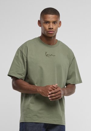 Tânăr cu păr scurt purtând un tricou larg verde măsliniu cu un logo discret scris, stând în picioare pe un fundal simplu, gri deschis.