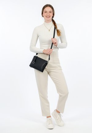 Černá kožená crossbody taška se stříbrnými detaily. Modelka má na sobě bílé žebrované rolák, béžové kalhoty a bílé tenisky.