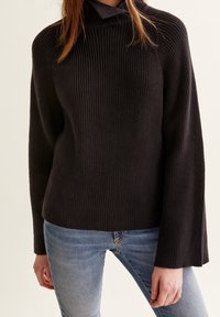 Svart stickad tröja med hög krage och ribbad textur, med långa trumpetärmar. Bärs över ljusblå jeans.