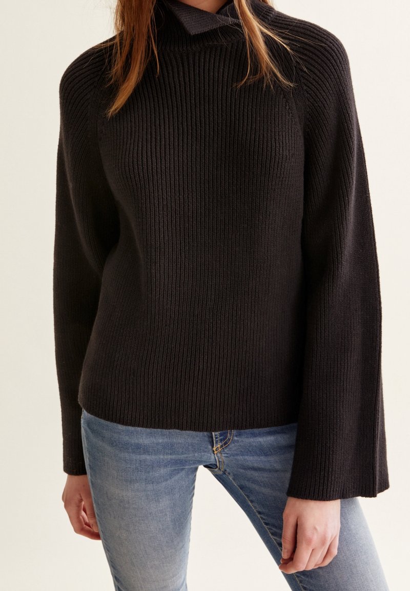 Svart stickad tröja med hög krage och ribbad textur, med långa trumpetärmar. Bärs över ljusblå jeans.