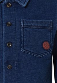 Jeansskjorta i mörkblått med knappstängning, krage och bröstficka. Har ett runt, brunt logopatch ovanför fickan.