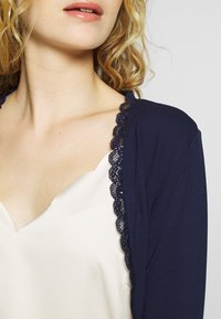 Marinblå cardigan med spetsdetaljer, lätt tyg, öppen framsidesdesign, tillsammans med en krämfärgad silkestopp. Närbild av halsringningen.