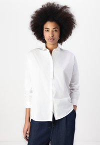 White cotton button-up shirt with long sleeves, a collar, and a relaxed fit, paired with dark denim pants. Simple and clean design.