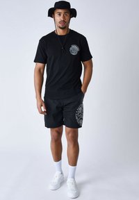 Camiseta negra con estampado gráfico en el pecho, combinada con pantalones cortos negros que tienen un diseño de logo blanco. Zapatillas blancas y calcetines deportivos.