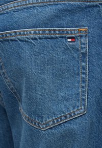 Τζιν σε μεσαίο μπλε χρώμα, με πίσω τσέπη που διαθέτει πορτοκαλί ραφές και μικρή ετικέτα με κόκκινο, λευκό και μπλε λογότυπο. Υφή στην επιφάνεια.