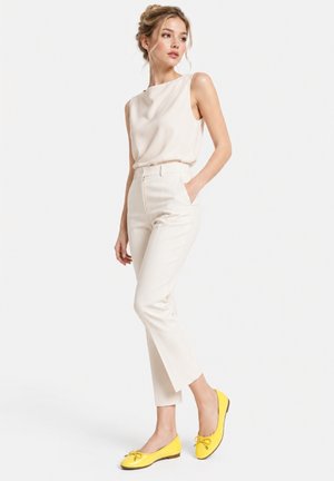 Woman with light hair in a sleeveless beige top and high-waisted cream pants stands with one hand in pocket, wearing bright yellow ballet flats.