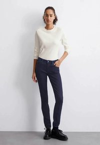 Sieviete stāv pie baltas sienas, tērpta baltā garās piedurknes topā, tumšās šaurajās biksēs un melnās šņorējamās kurpēs.