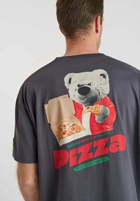 Man in een donkergrijs shirt met een afbeelding van een beer in een rode jas die pizzastukken vasthoudt, met het woord "Pizza" in grote rode letters.