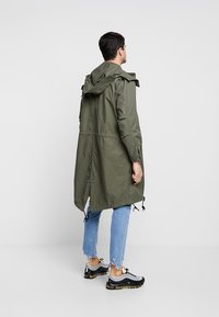 Parka verde oliva con cappuccio, cordini regolabili e un orlo posteriore più lungo. Abbinato a jeans blu strappati e sneakers sportive.